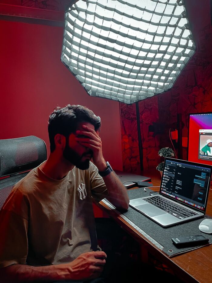 A man holding his head in a dimly lit room with a laptop, reflecting simple joys that are mainstream.