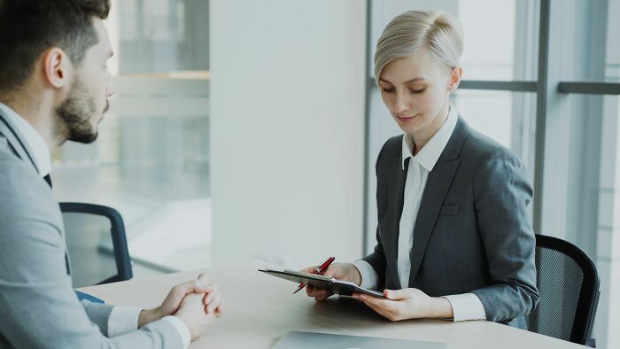 A manager in a grey suit interviews a male candidate, suggesting corporate hiring scandals.