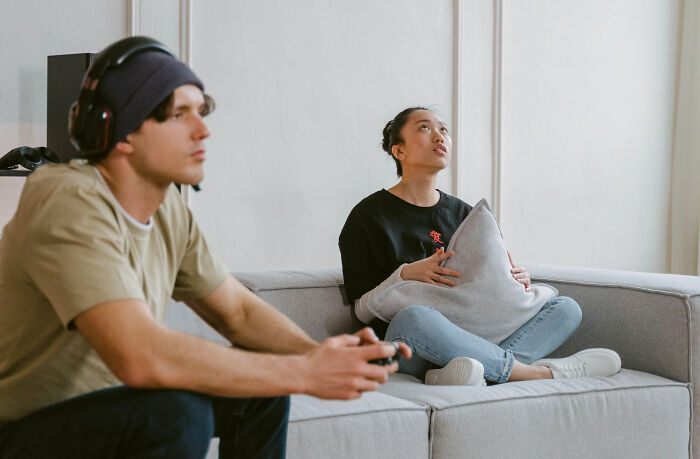 A woman sits on a couch, holding a pillow, looking bored while a man plays video games, hinting at dating red flags.