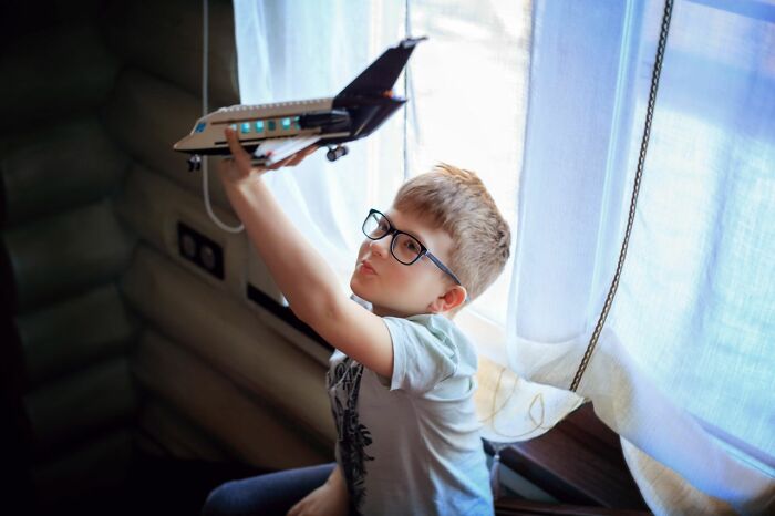 A young boy wearing glasses plays with a toy airplane, dreaming of future airline seating lottery adventures.