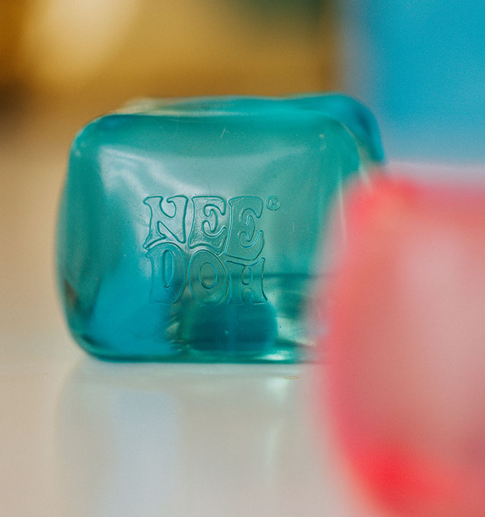 Close-up of a NeeDoh squishy toy in teal with a blurred pink squishy toy in the background, showing brand for squishy toys. Close-up of a NeeDoh squishy toy in teal with a blurred pink squishy toy in the background, showing brand for squishy toys.
