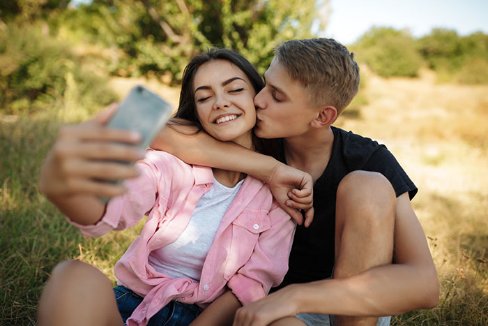 Teen boyfriend kissing his girlfriend as she takes a selfie, reflecting on a petty revenge plot. Teen boyfriend kissing his girlfriend as she takes a selfie, reflecting on a petty revenge plot.