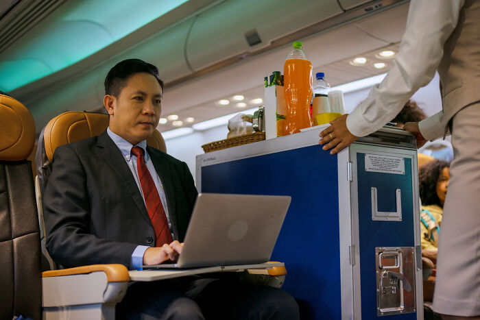 A passenger on a flight working on his laptop while a flight attendant serves drinks, showing things passengers do.