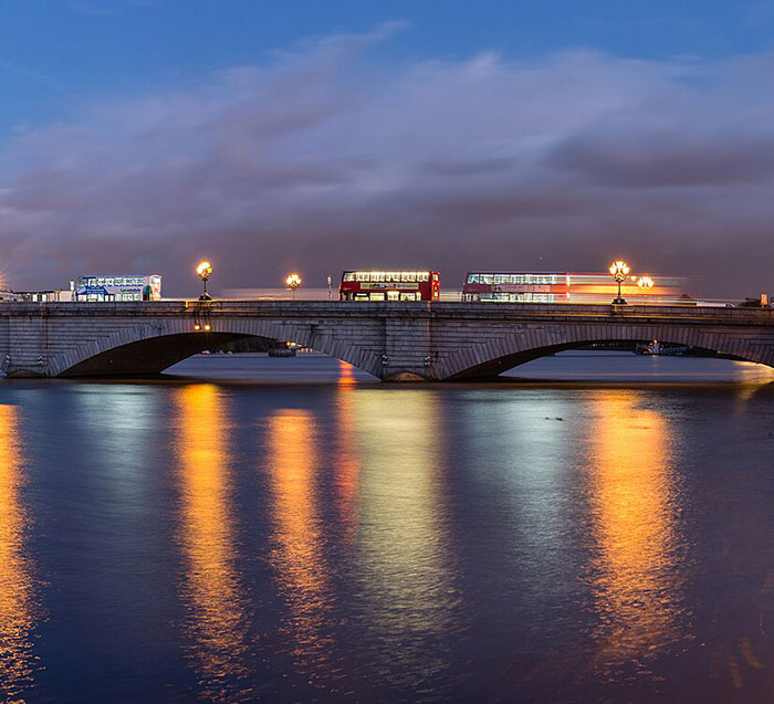London bridge at night, with red double-decker buses in motion, reflecting light on the water.
