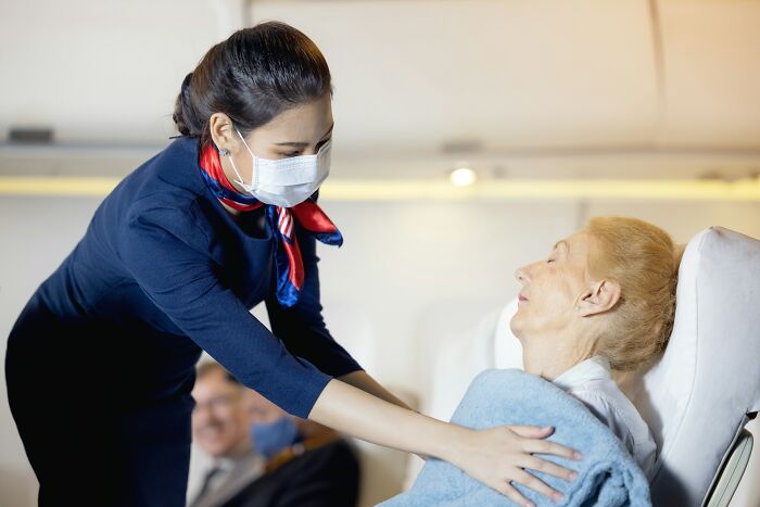 Cabin crew member wearing a mask assists an elderly female passenger on a plane, showing care in dealing with situations.