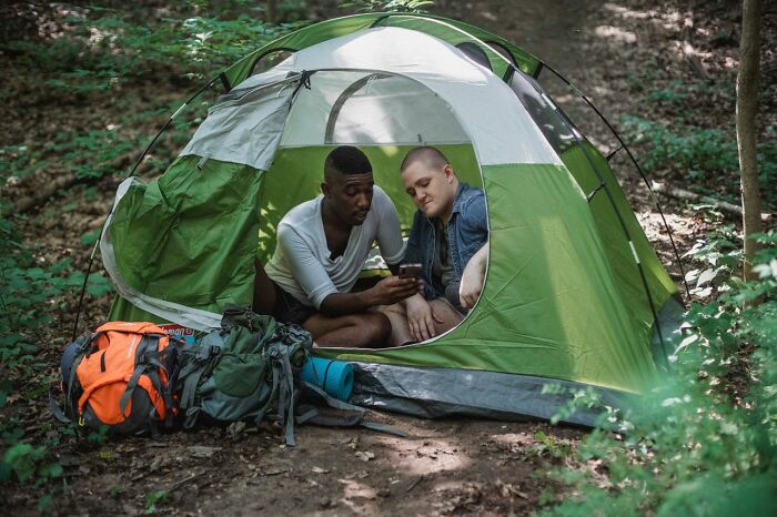 Two friends camping in a green tent looking at a phone, sharing relationship revenge stories.