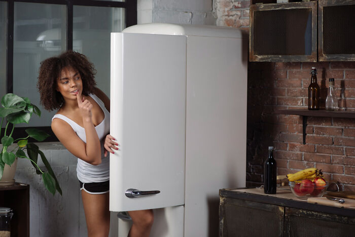 A mischievous woman peeks out from behind an open white refrigerator, planning relationship revenge.