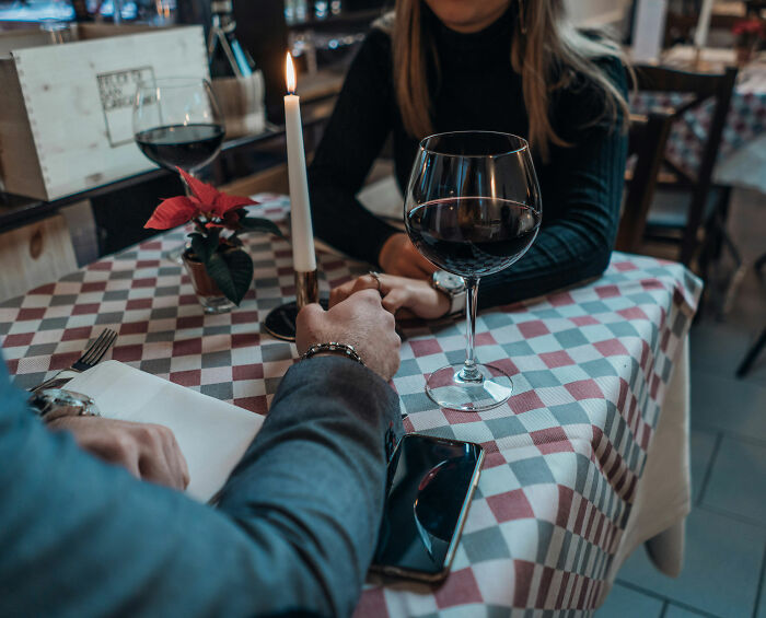 A couple holding hands across a checkered table, with wine glasses and a candle, representing a dating profiles first date. A couple holding hands across a checkered table, with wine glasses and a candle, representing a dating profiles first date.