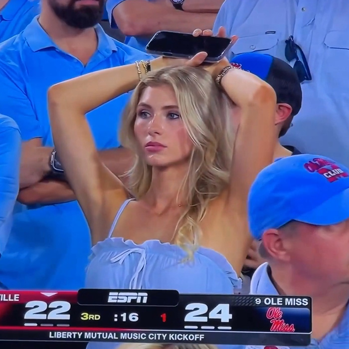 An attractive woman in football stands looks up, holding a phone above her head. She went viral for being spotted.