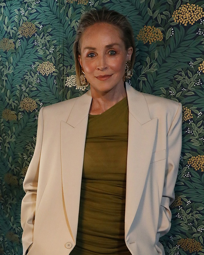 A-list actress Sharon Stone smiling in a beige blazer and olive dress against a floral background.