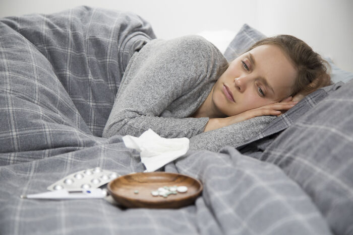 A woman looks unwell in bed with tissues and medication nearby, showing the reality of sickness and illness.