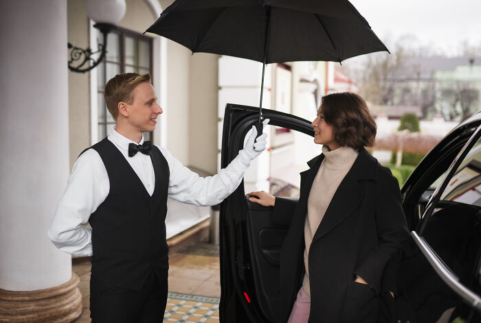 A doorman in a tuxedo holding an umbrella for a woman exiting a luxury car, reflecting services wealthy people buy.