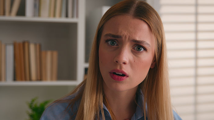 A young woman with a surprised expression, wearing a denim shirt, with bookshelves in the background.