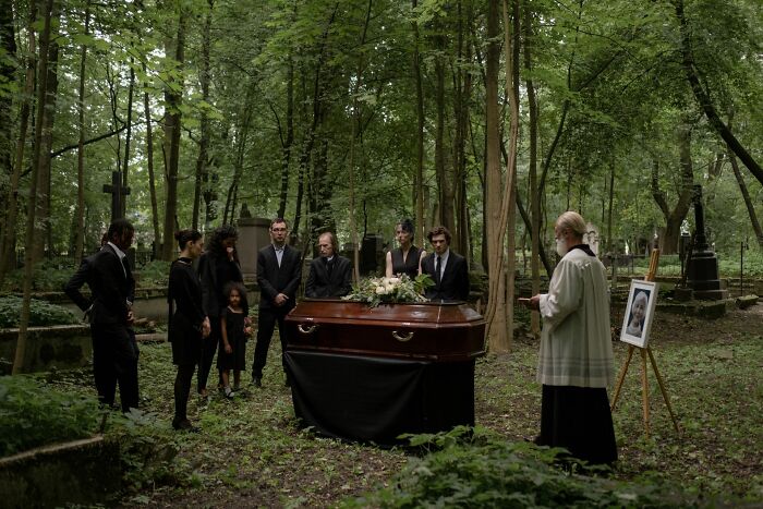 A group of people standing around a casket in a forest, an unsettling scene with hidden stories.
