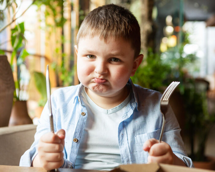 A young boy with a pouting face, holding a fork and knife, looking displeased. Unsettling stories from small towns.