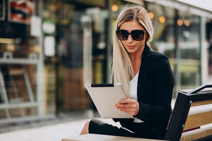 A blonde woman in sunglasses and a black blazer sits on a bench, looking at a tablet, hinting at unsettling small town stories.