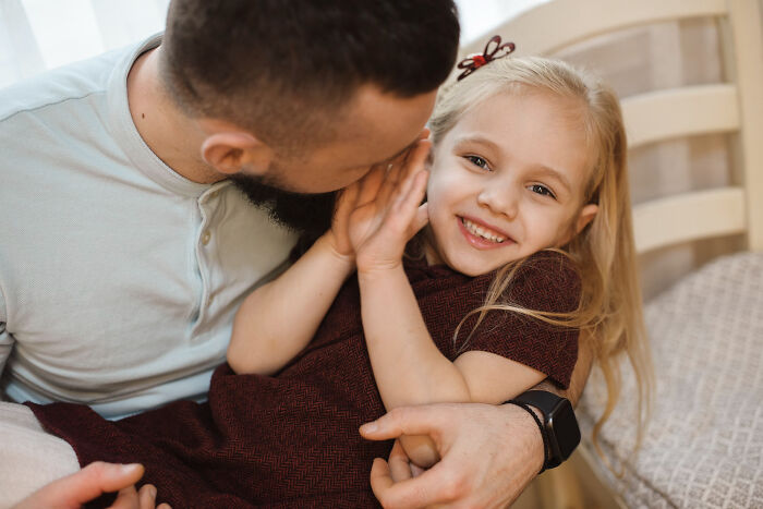 A smiling young girl with blonde hair in a red dress is held by a bearded man who whispers in her ear, sharing small town secrets.
