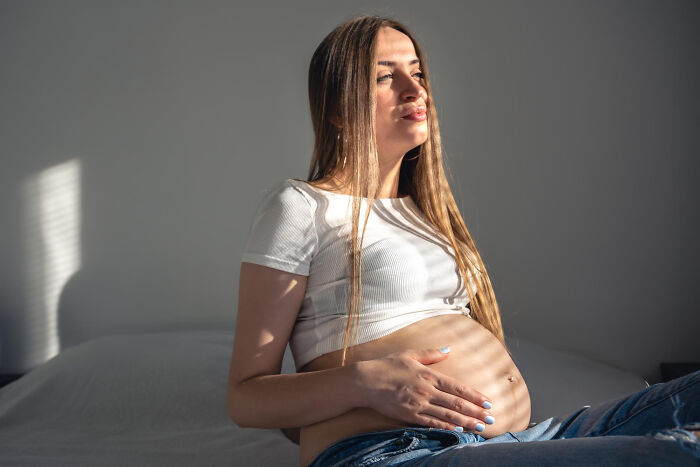 A pregnant woman holding her belly, sitting by a window with sunlit stripes. Unsettling stories from small towns often involve secrets.