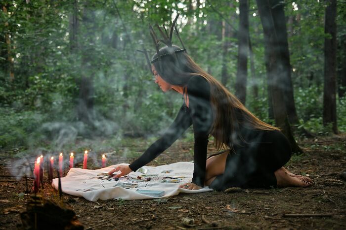 Woman in a dark dress with a crown, performing a ritual with candles and objects in a misty forest, evoking shocking secrets.