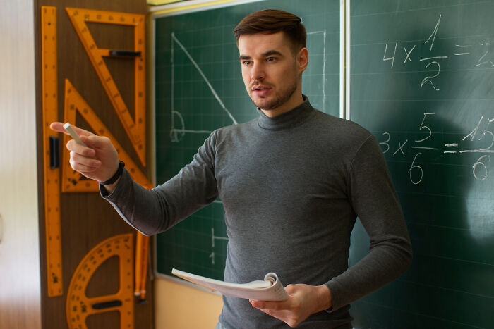 A teacher explains, holding chalk and paper, in a classroom with mathematical equations on a green board, revealing small town secrets.
