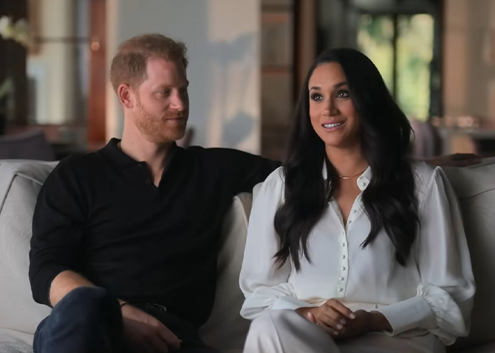 Meghan Markle and Prince Harry sitting on a sofa, looking at the camera. They are discussing their UK return. Meghan Markle and Prince Harry sitting on a sofa, looking at the camera. They are discussing their UK return.