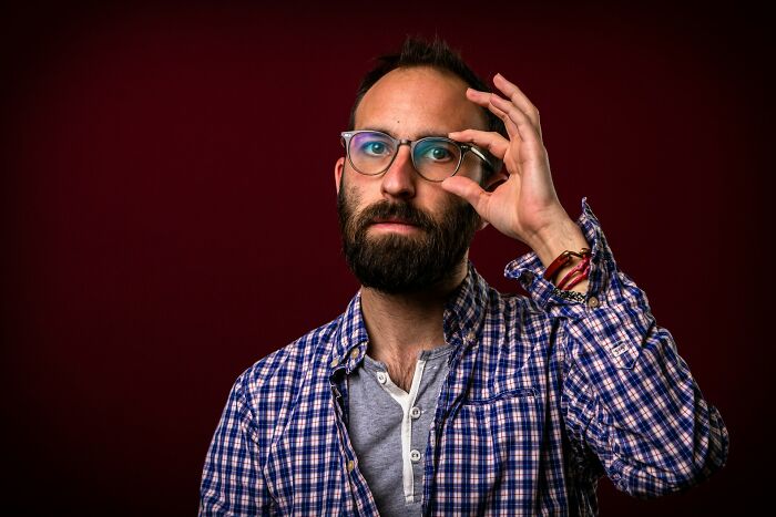 A bearded male medical staff member adjusting his glasses on a dark red background