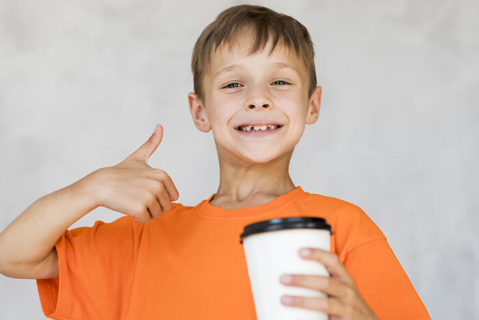 A smiling boy in an orange shirt giving a thumbs up while holding a cup, a weird sight at someone's home.