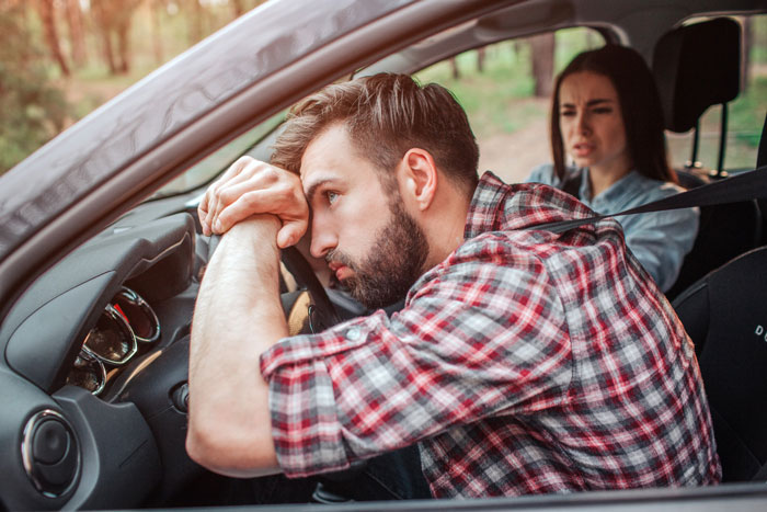 Upset man with beard resting his head on the steering wheel, with a disapproving woman in the background, showing the impact of parents ruining a boyfriend's reputation. Upset man with beard resting his head on the steering wheel, with a disapproving woman in the background, showing the impact of parents ruining a boyfriend's reputation.