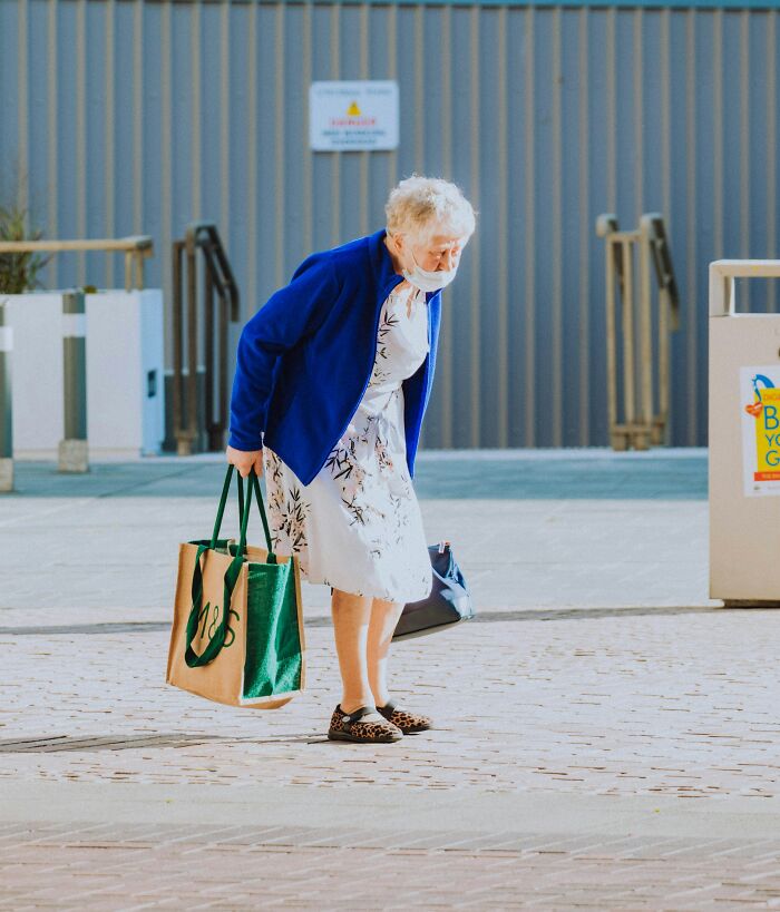 An elderly woman wearing a face mask and a blue cardigan, leaning over while carrying shopping bags, suggesting physical pain reactions.