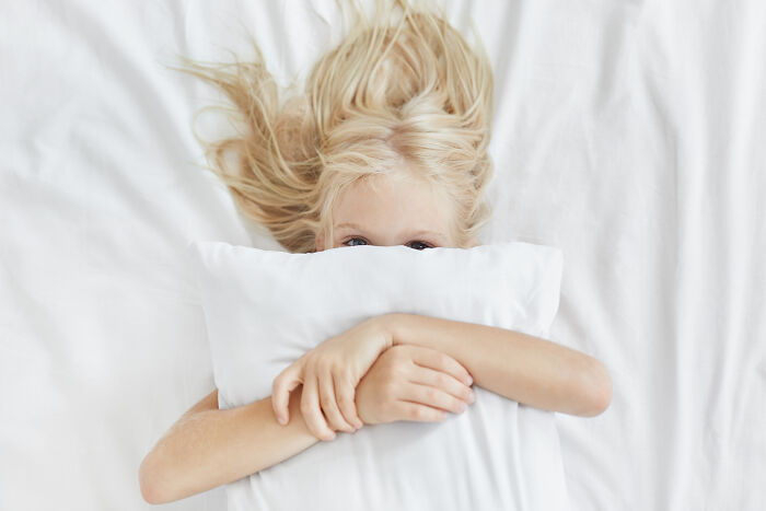 A blonde child with blue eyes peeking over a white pillow, depicting childhood beliefs.