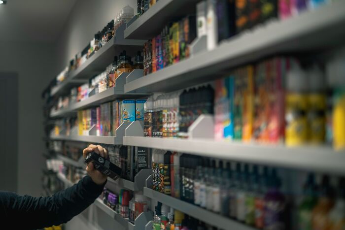 Hand selecting a vaping product from a shelf full of various e-liquids and vape devices, related to a vaping habit. Hand selecting a vaping product from a shelf full of various e-liquids and vape devices, related to a vaping habit.