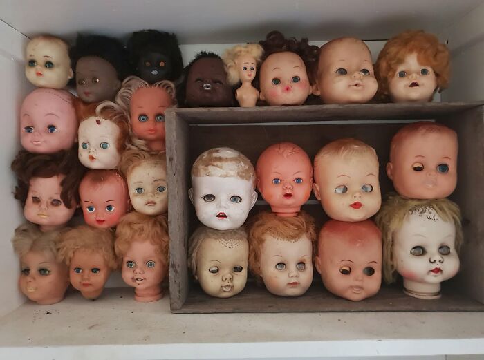 A collection of unhinged secondhand doll heads, some with missing eyes or discolored skin, arranged in a wooden crate.