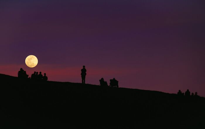 Silhouettes of people on a hill watching a full moon rise, a beautiful scene illustrating cool facts about the universe.