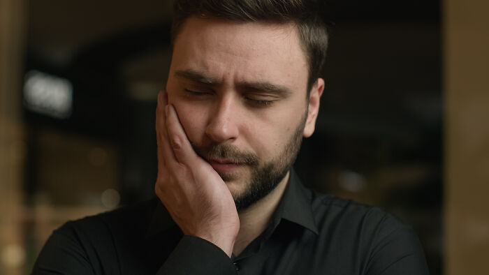 A man with a beard, eyes closed, holding his face in distress, experiencing extreme illness.