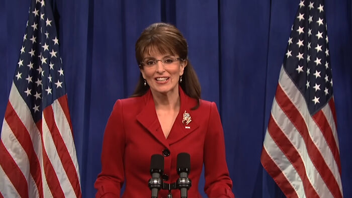 Tina Fey impersonating Sarah Palin behind a podium flanked by American flags
