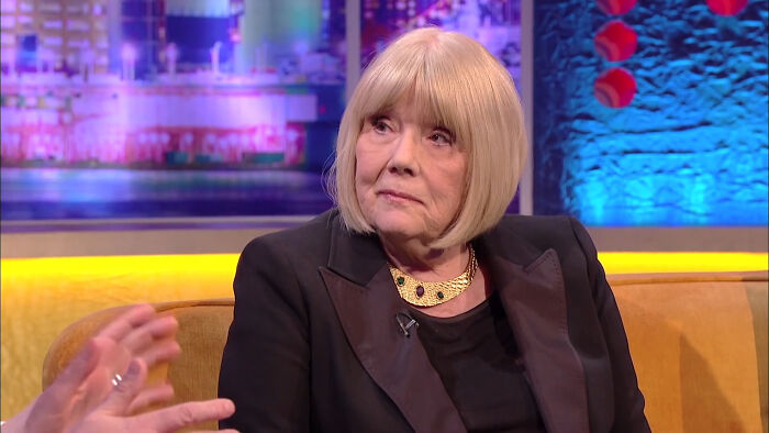 A stage actress with blonde bob hair, wearing a black blazer and gold necklace, on a talk show.