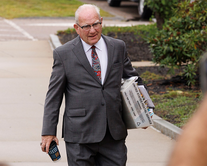 An older man walking outdoors, holding a box of mail, related to the Lindsay Clancy wheelchair sticker incident. An older man walking outdoors, holding a box of mail, related to the Lindsay Clancy wheelchair sticker incident.