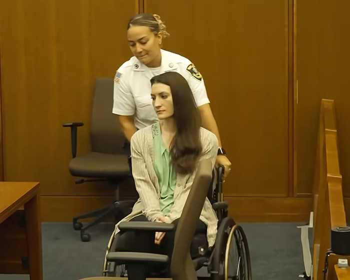 Lindsay Clancy in a wheelchair, being pushed by a corrections officer, with an odd sticker visible on the wheelchair. Lindsay Clancy in a wheelchair, being pushed by a corrections officer, with an odd sticker visible on the wheelchair.