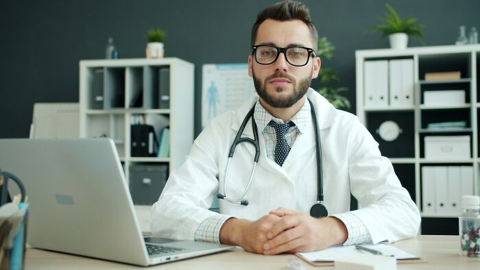 A male medical staff doctor sitting at his desk with a laptop and looking forward with a stethoscope