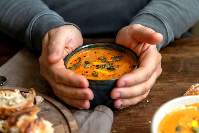 Person holding a bowl of pumpkin soup garnished with seeds and herbs