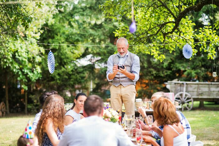 A happy older man standing at an outdoor table with guests, celebrating a wild wedding turn in a garden.