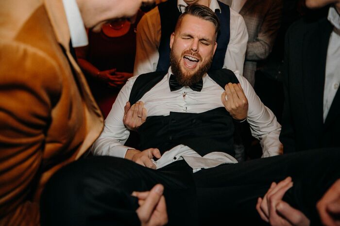 A man in a tuxedo is held up, laughing joyfully, celebrating a wild wedding turn with friends.