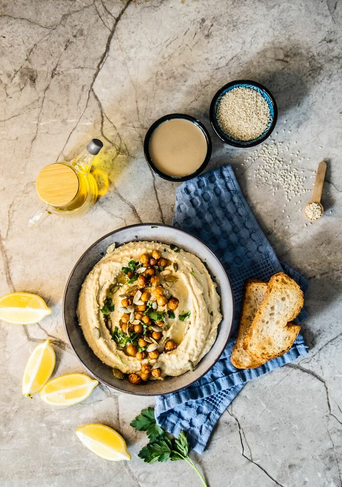 A bowl of hummus topped with chickpeas and seeds, surrounded by lemons and toast, offering a healthy snack.