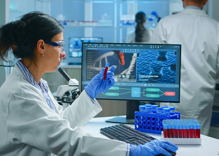 A woman in a lab coat and safety glasses holding a test tube with a DNA test result on a computer screen, symbolizing Ancestry DNA Test.