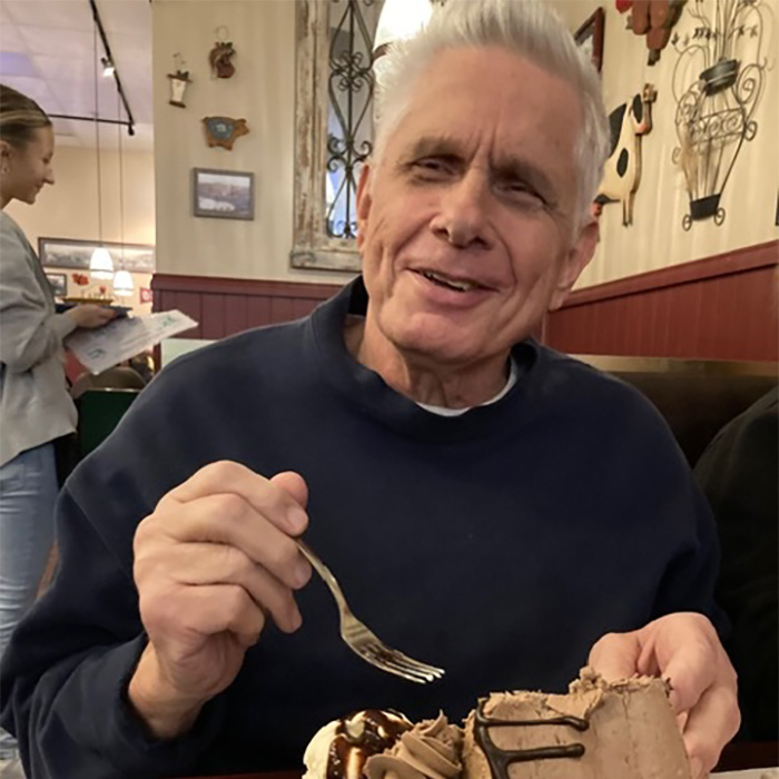 Husband eating dessert, smiling in a restaurant. His wife was arrested for leaving him on the toilet.