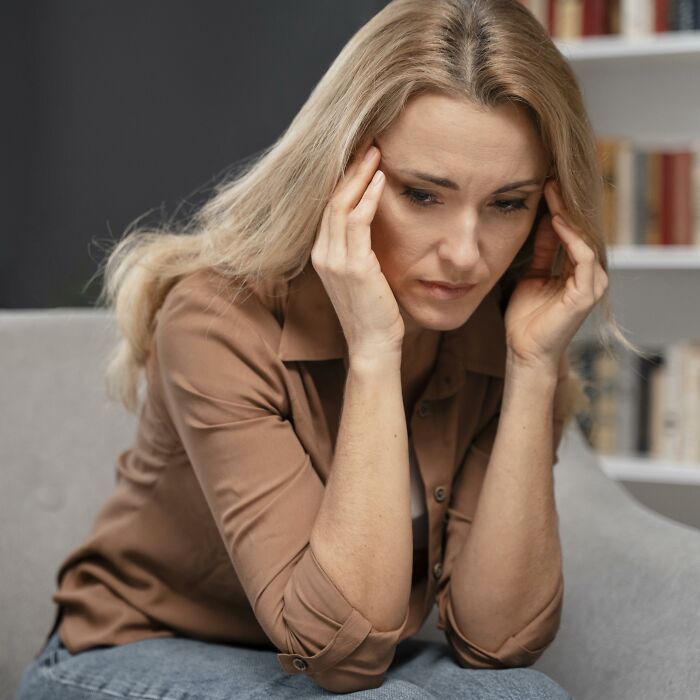 A woman with blonde hair looking upset, holding her temples, sitting on a sofa. This image depicts the emotional impact of men's lies.