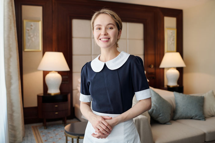 A smiling housekeeper in a uniform stands in a luxurious hotel room, a service wealthy people buy.