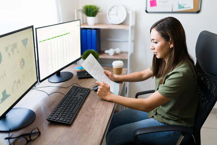 A professional woman reviewing documents at a desk with multiple monitors, uncovering industry secrets.