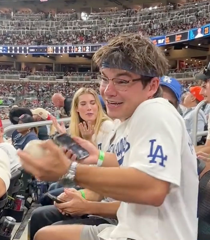 A happy Dodgers fan holding a baseball in the stadium stands, smiling and talking.