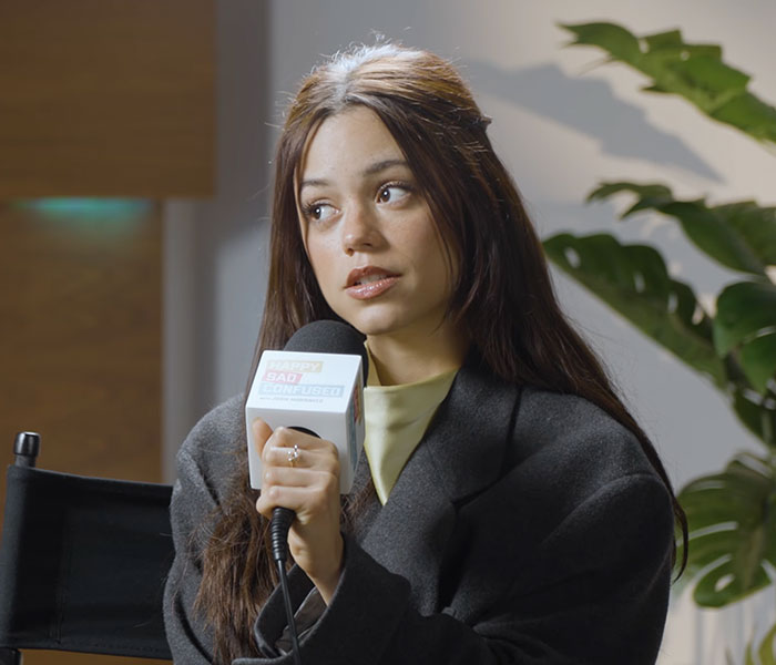 Jenna Ortega holding a Happy Sad Confused podcast microphone during an interview. Jenna Ortega holding a Happy Sad Confused podcast microphone during an interview.
